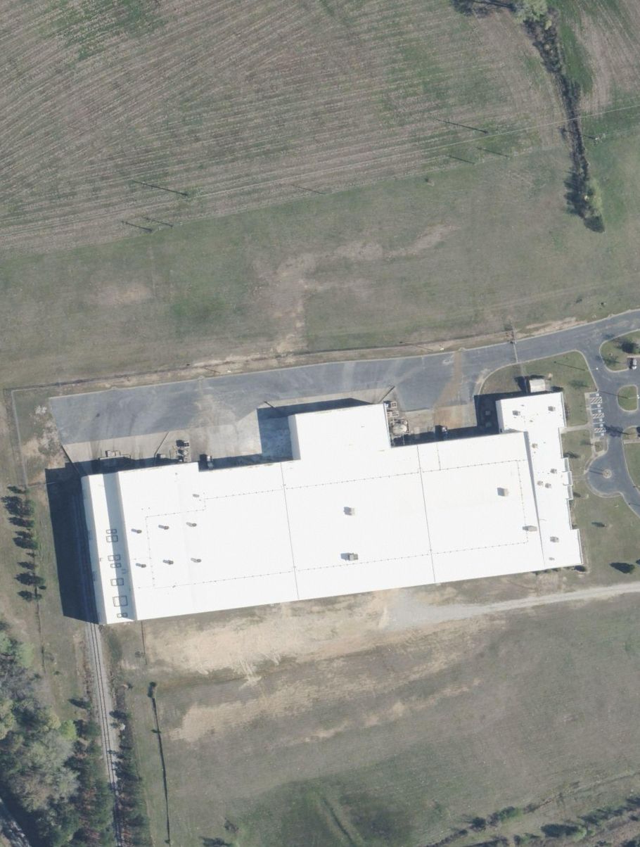 Aerial view of a large white industrial building with a flat roof and surrounding parking lot and fields.