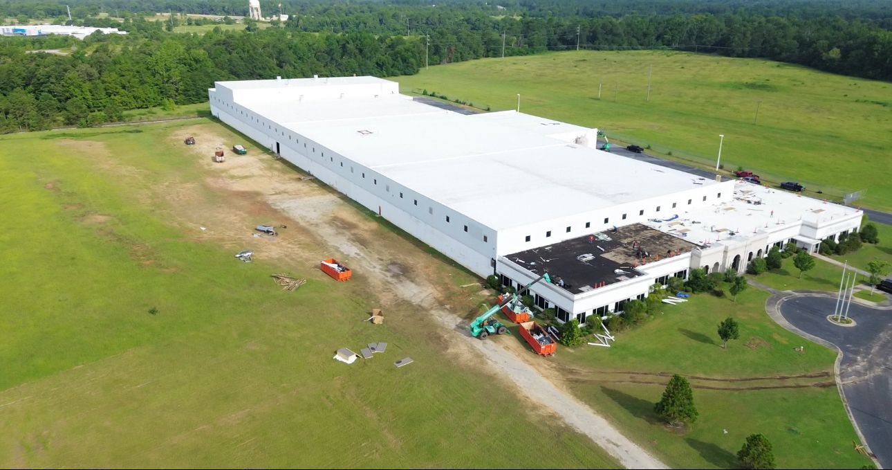 Large white industrial building on a green field with trees in the background. Construction equipment visible.