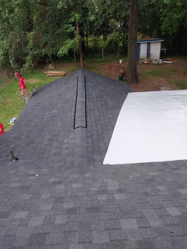 Dark gray shingled roof with a white flat roof section; trees in the background, a small building.