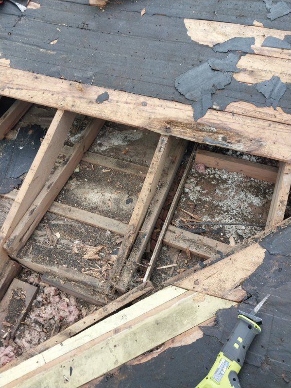 Overhead view of a roof under construction; exposed wooden rafters, partially removed roofing, and a reciprocating saw.
