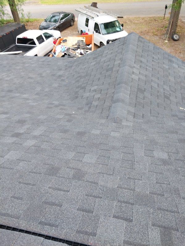 Dark gray asphalt shingle roof. White truck, car, and equipment in the background, near a road.