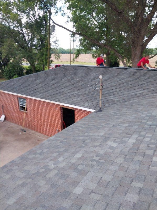 Two roofers in red shirts on a dark shingled roof, next to a red brick building, working.