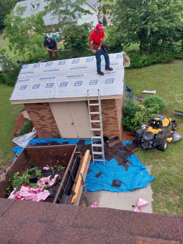 Roofers on a small building, replacing shingles. One on roof, another on ground, surrounded by debris.