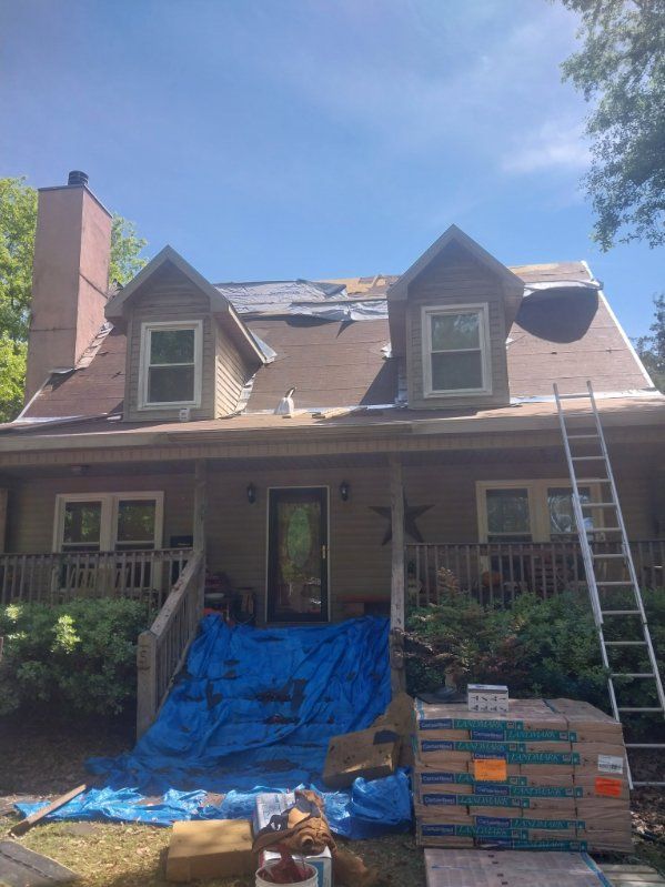 House with partially removed roof, blue tarp on porch, ladder, and bundles of shingles.