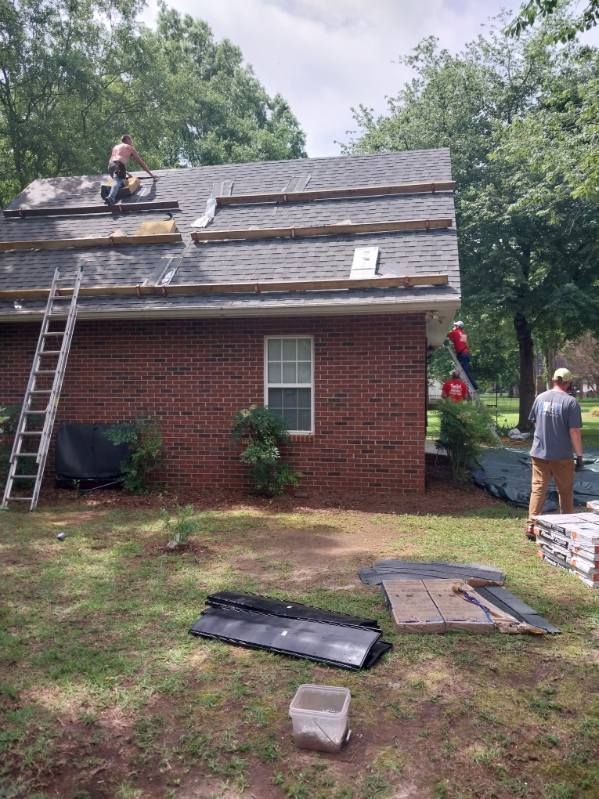 Roofers working on a brick house roof. Tools and materials are on the ground and roof. Trees in the background.