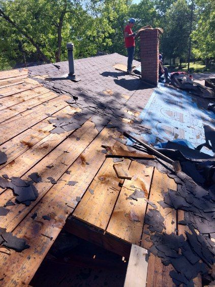 Roofer removing old shingles from a wooden roof, near a brick chimney, on a sunny day.