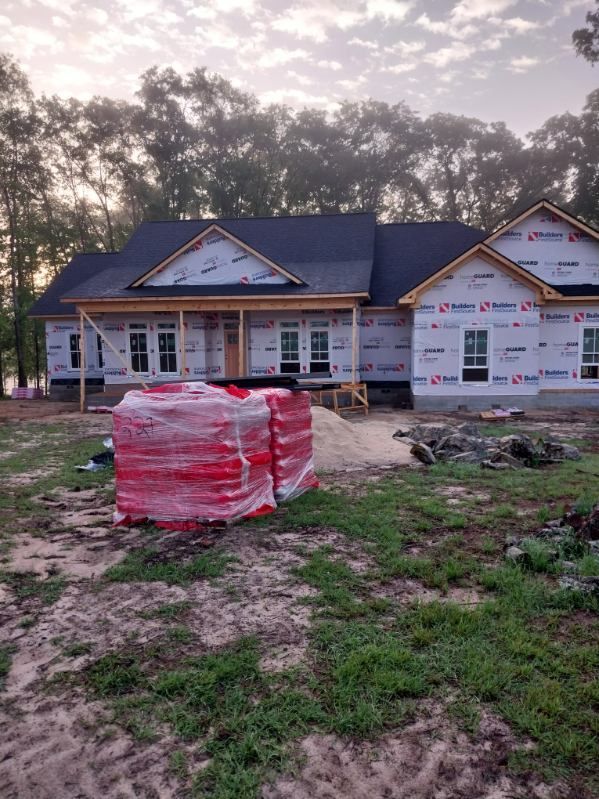 House under construction with stacks of red bricks in front. Wrapped in white sheathing, surrounded by trees.