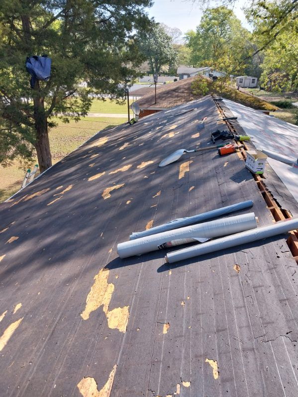 Roof with exposed underlayment and tools, trees, and sky in the background.