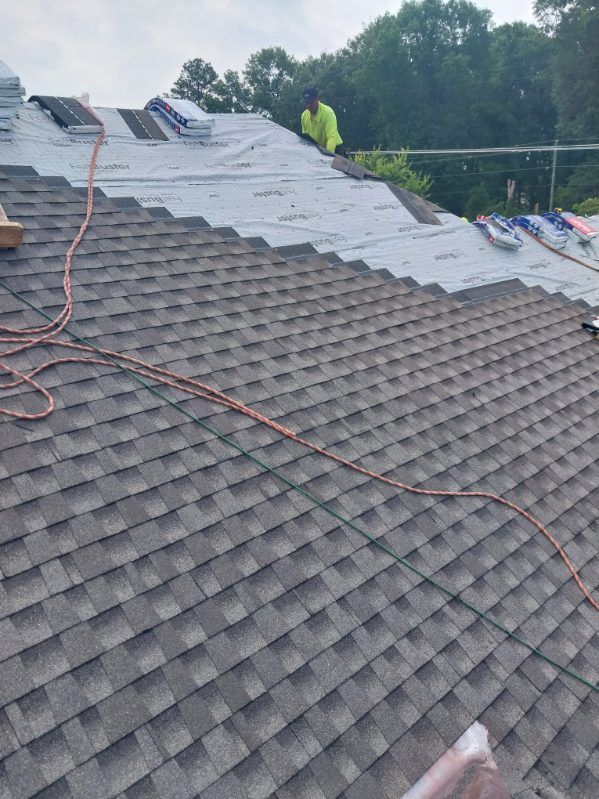 Roofer on a house roof installing dark gray shingles, with underlayment visible and supplies nearby.