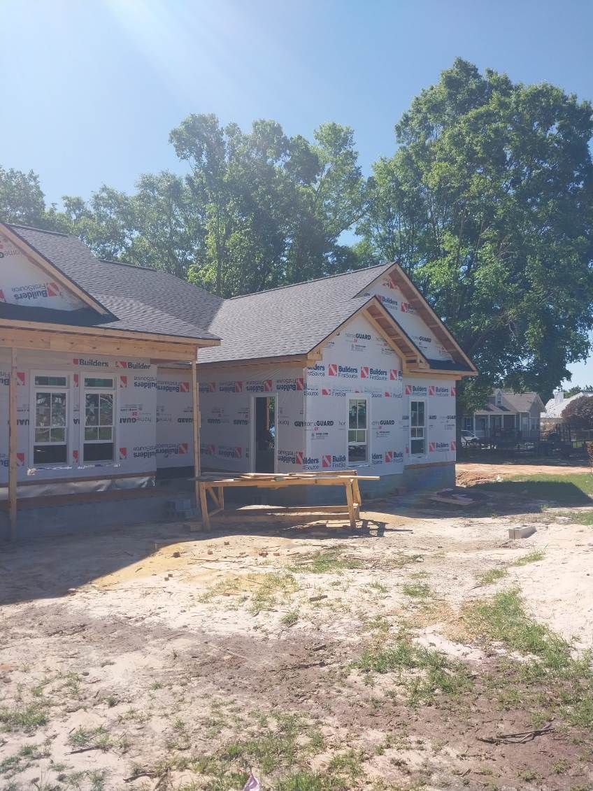 Construction site of a house with exposed framing and wrap, blue sky, and trees.