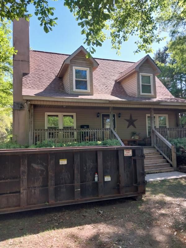 House with brown roof and siding, two dormers, a front porch, and a large dumpster in the foreground.
