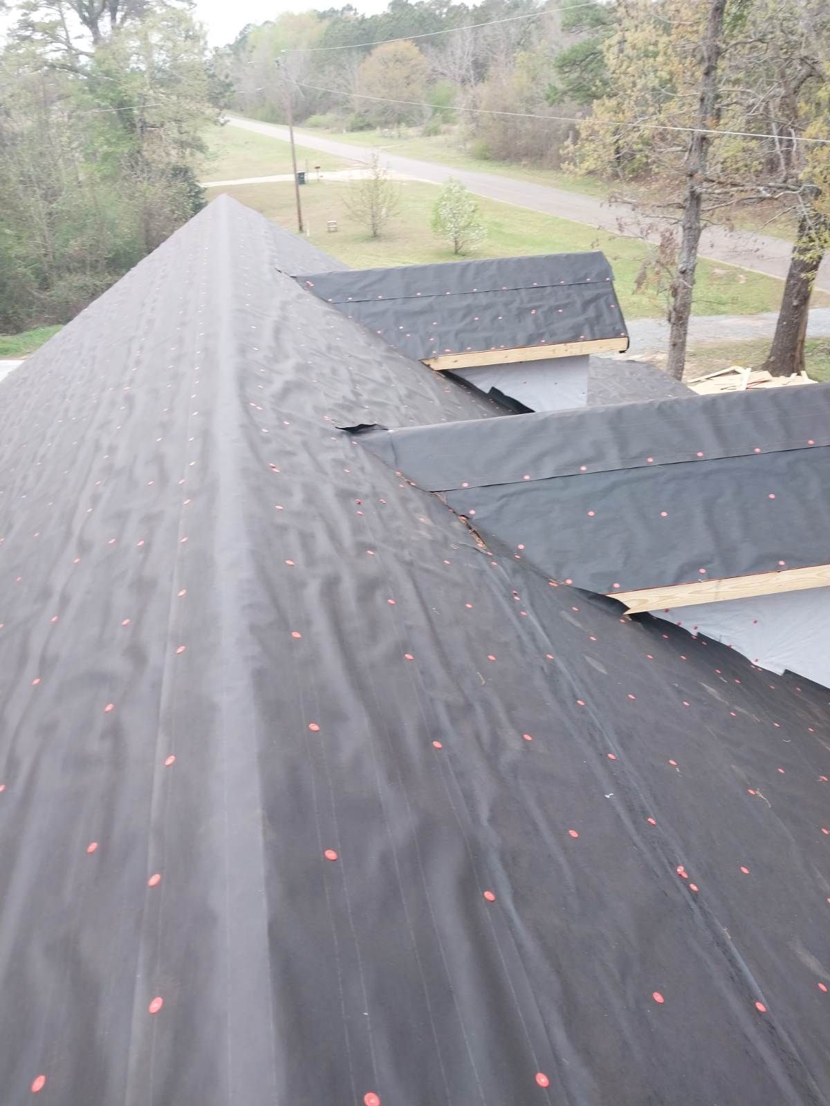 Black roofing material being installed on a roof with a view of a road and trees in the background.
