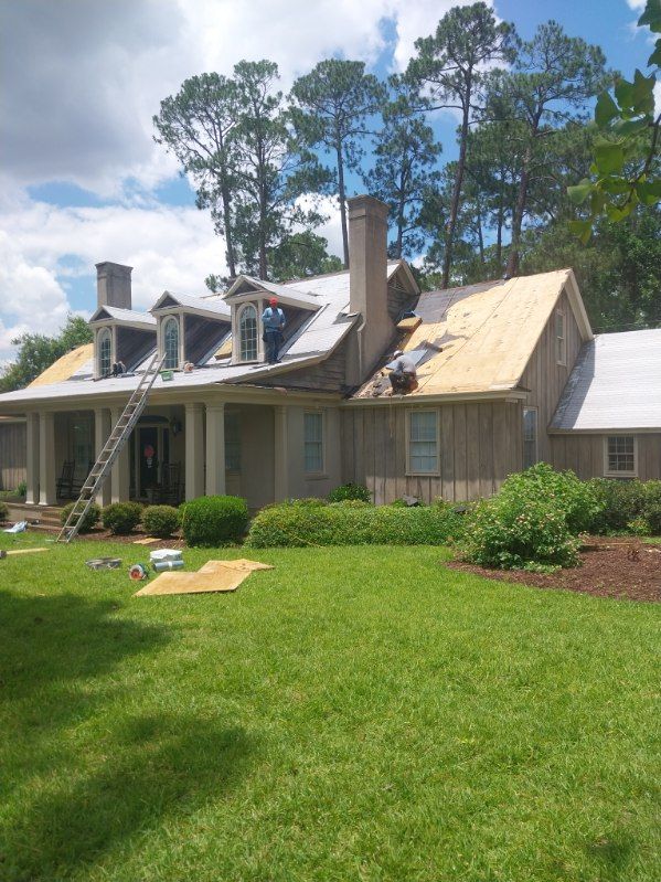Roofers working on a house with a partly removed roof; ladder, green grass, trees, and blue sky in the background.