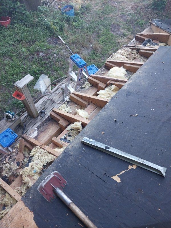 Roof construction: exposed wooden beams, insulation, and tools on a flat roof, with a garden in the background.