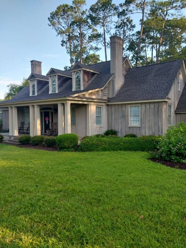 A light-colored house with a porch, white columns, and dormer windows, surrounded by green grass and trees.