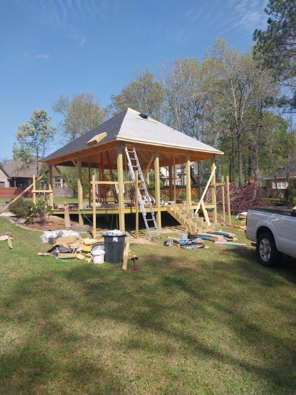 Gazebo under construction, wood frame with metal roof. Ladder propped up. Outdoors, sunny day.