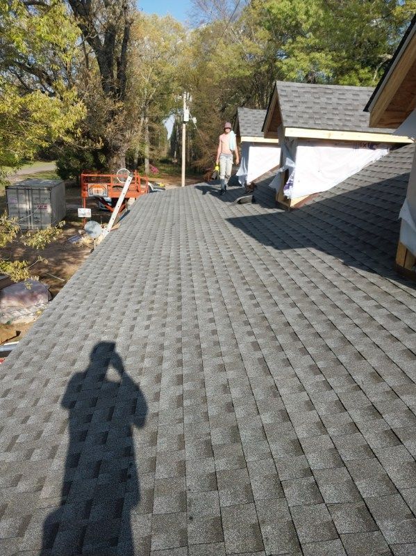 A person stands on a shingled roof, trees in the background. Construction site.