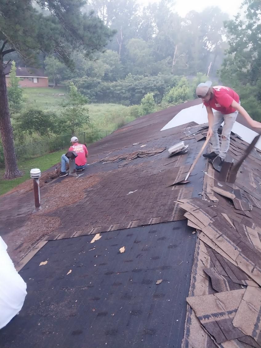 Two people remove shingles from a roof. The person on the right uses a scraper. Overcast day.