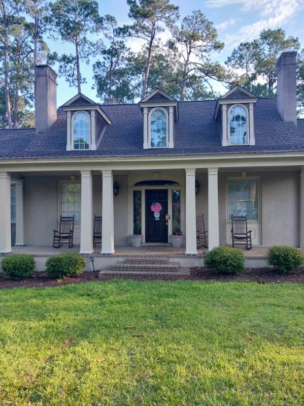 Gray house with white columns, three dormers, and a front porch. Two rocking chairs. Green lawn.