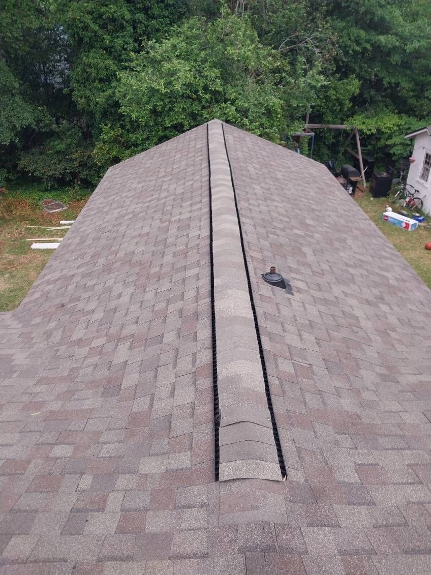 View of a roof with brown shingles, a central valley, and a backdrop of trees.