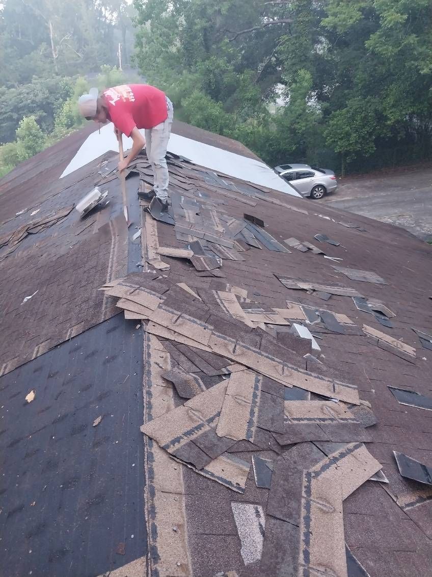 Person in red shirt on a damaged roof, removing shingles. Outdoors, trees in background, car parked.