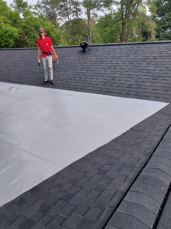 Person standing on roof, part shingled, part coated in white sealant. Trees in background.