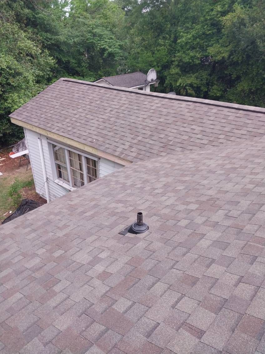 Brown asphalt shingle roof on a house with a black vent stack against a backdrop of trees.