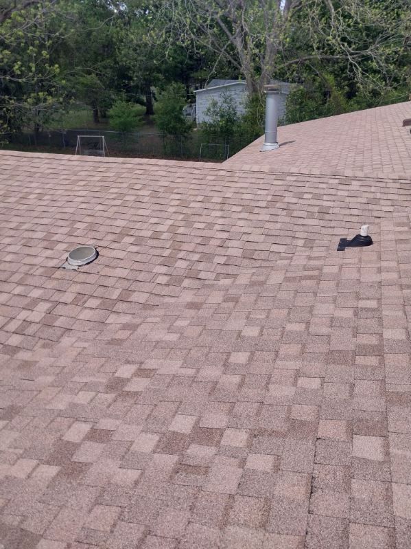Brown asphalt shingle roof with vents and a chimney against a background of trees.