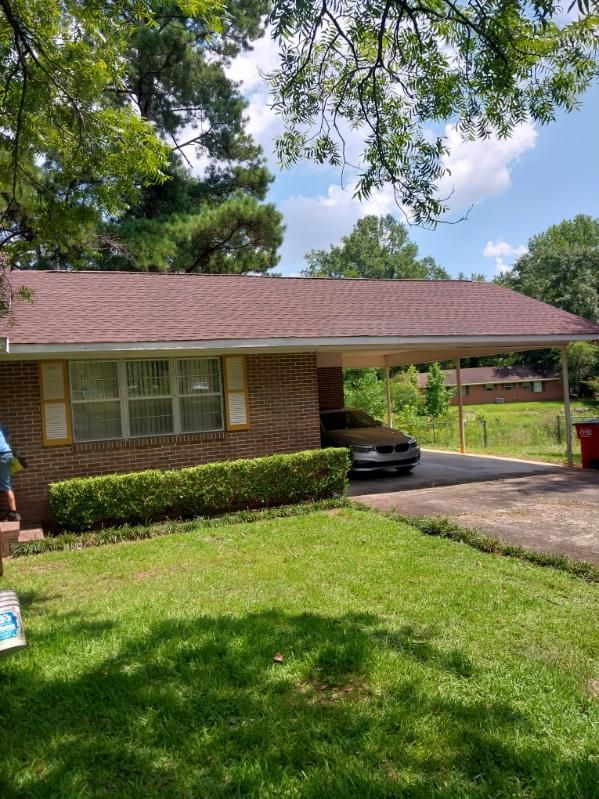 A brick house with a brown roof and carport. A car is parked in the carport, with bushes and green lawn in front.