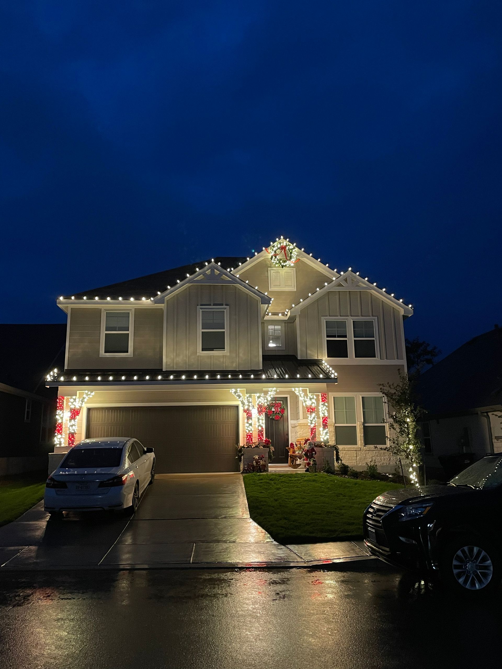 A house decorated with white Christmas lights; cars parked in the driveway; wet road reflecting lights; dark blue sky.