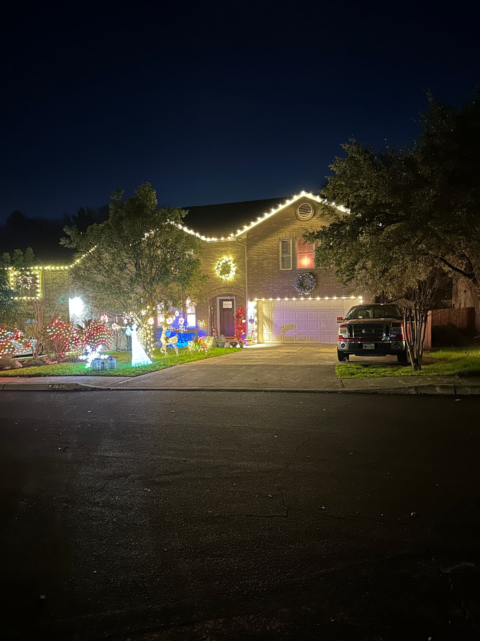 House at night decorated with white Christmas lights, wreaths, and figures, with a truck parked in the driveway.
