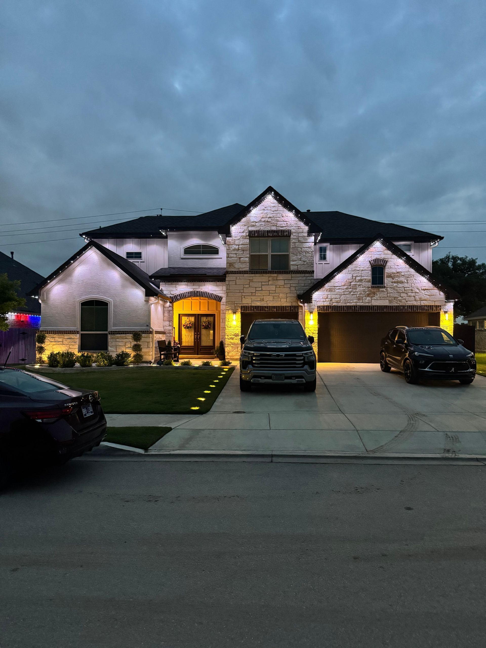 Two-story house at dusk with accent lighting, cars in driveway, and a cloudy sky.