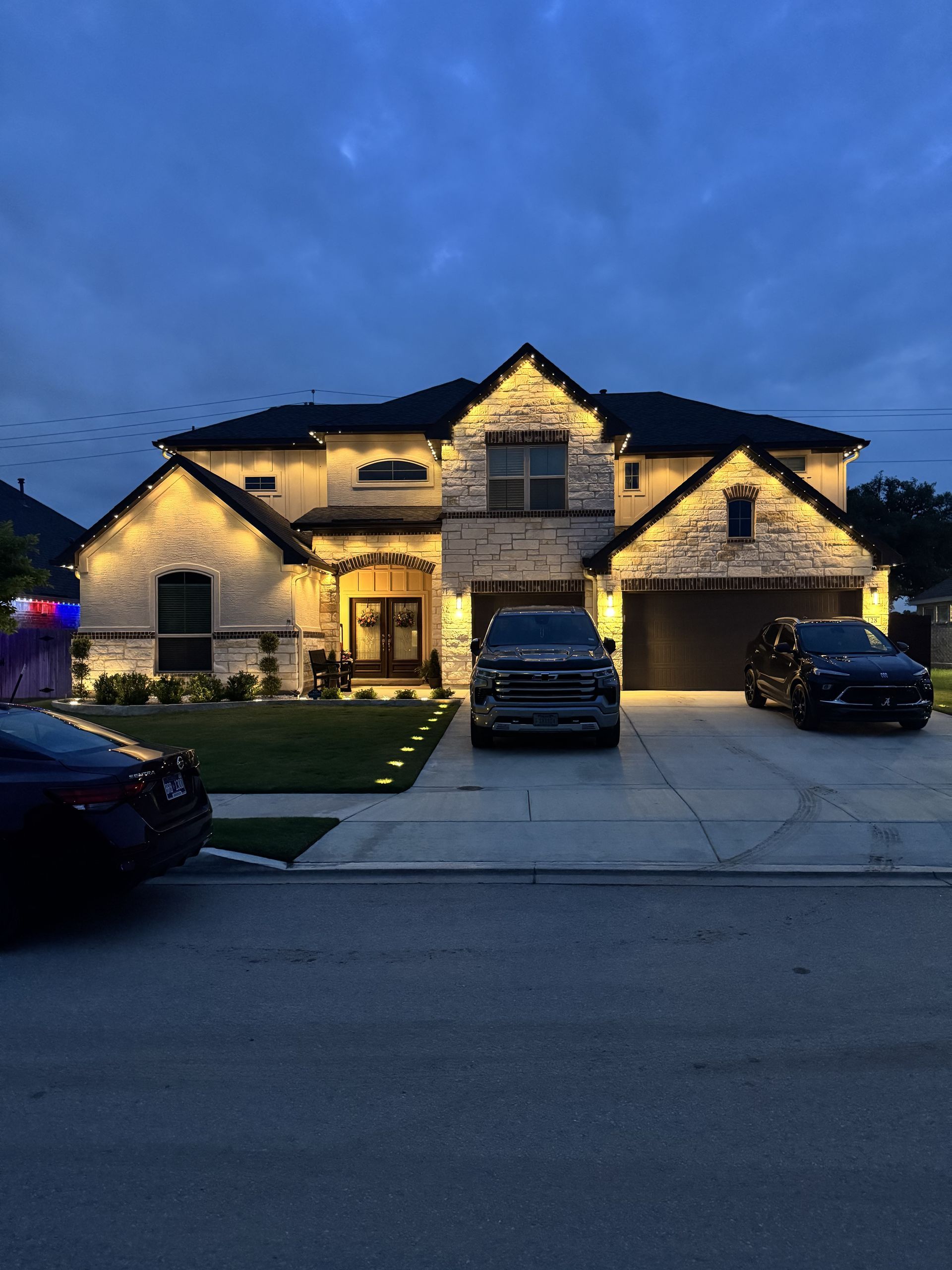 Two-story house with outdoor lighting at dusk; cars in driveway; blue sky.