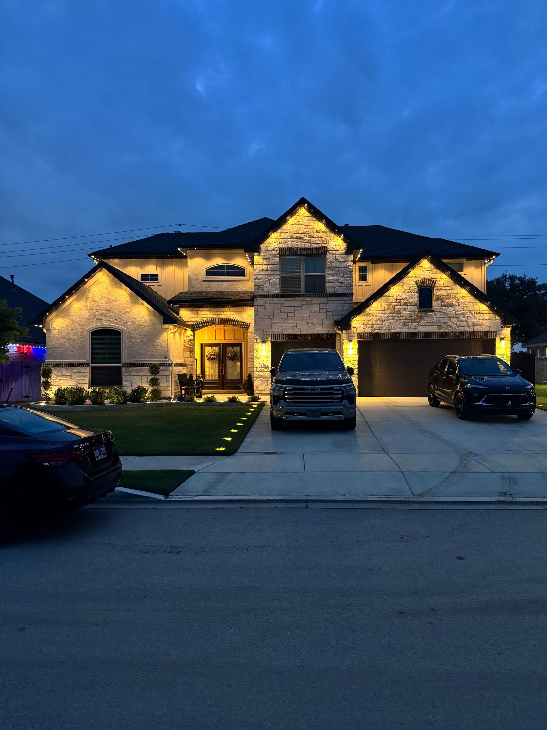 House exterior illuminated at dusk; stone facade, two-car garage, driveway with two vehicles parked out front.