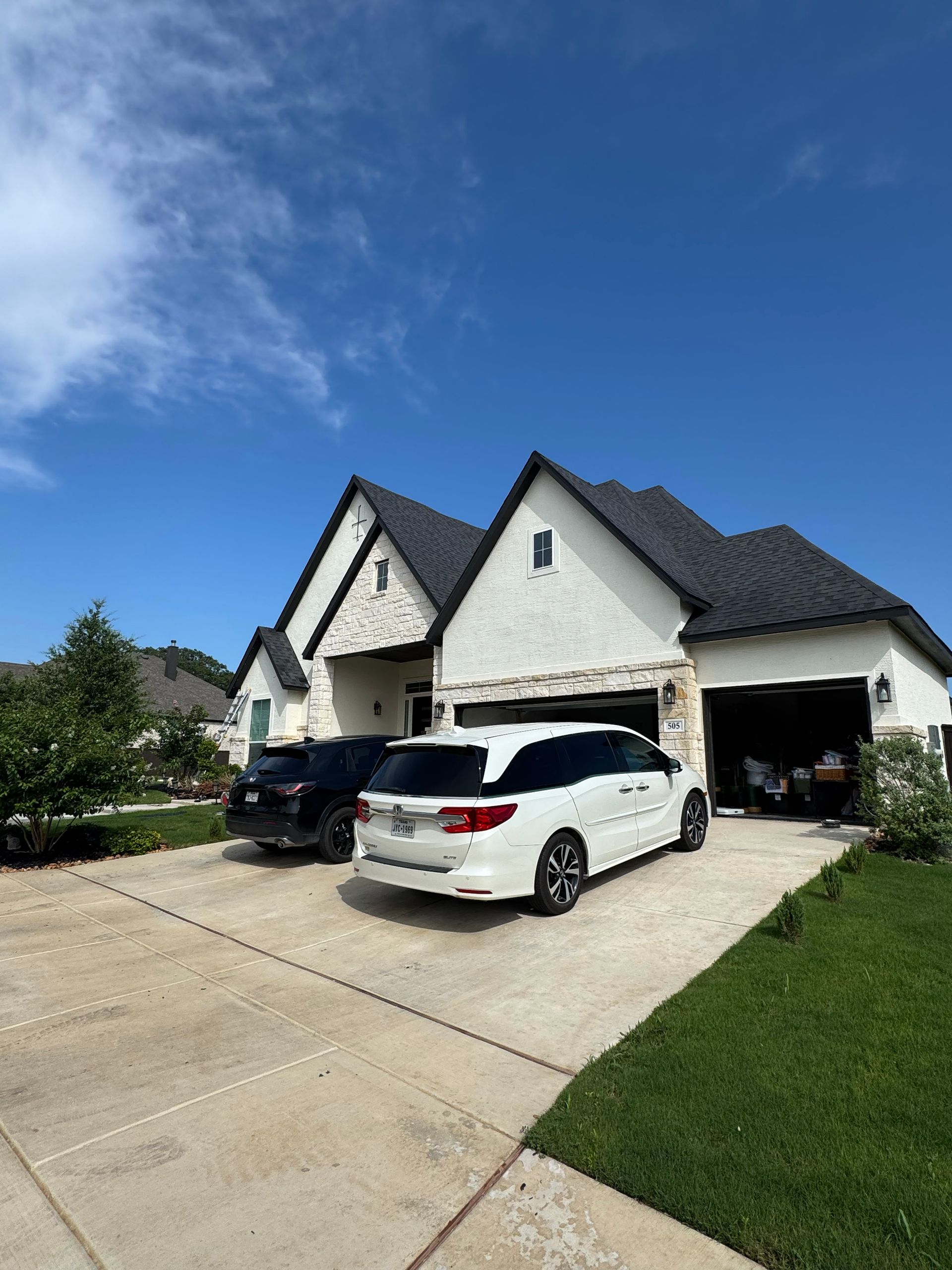 A white house with black roof, a black car, and a white minivan parked on the driveway on a sunny day.