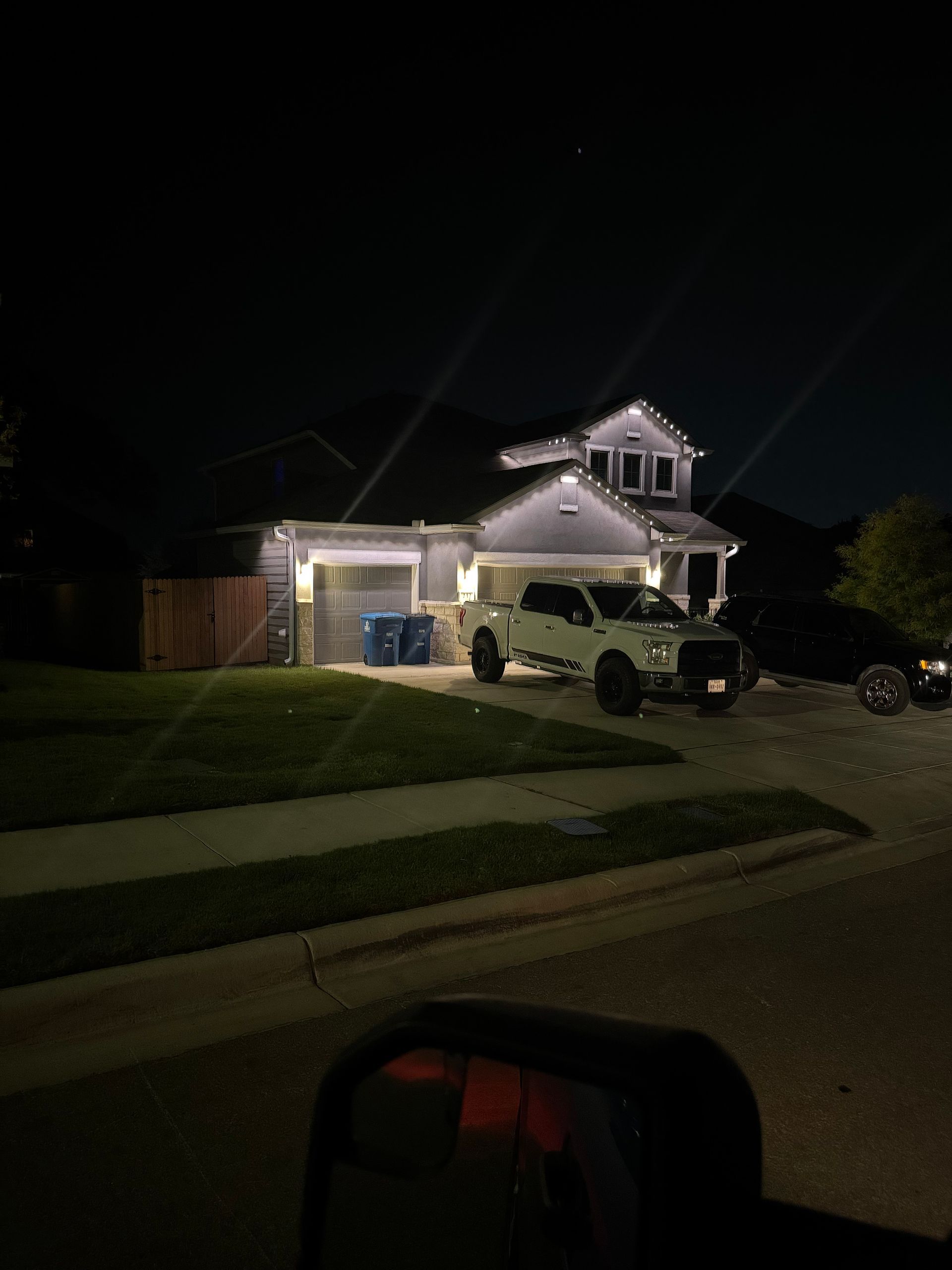 Night scene: a house with lit-up exterior, a white truck in the driveway, and a black car parked next to it.