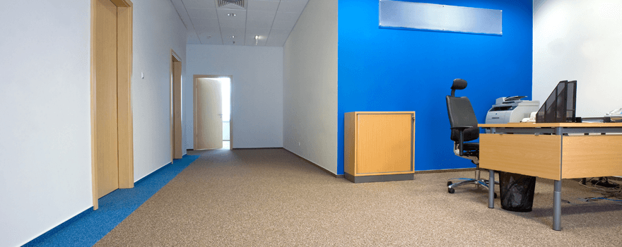 A wooden desk and black chair in a corridor recess, on light brown carpet, with a bright blue feature wall behind the desk