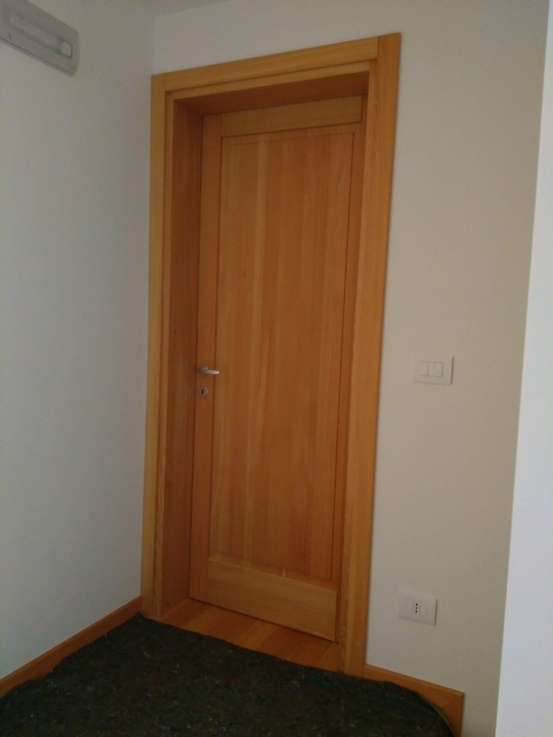 Una porta di legno si trova in un corridoio, accanto a un muro bianco.