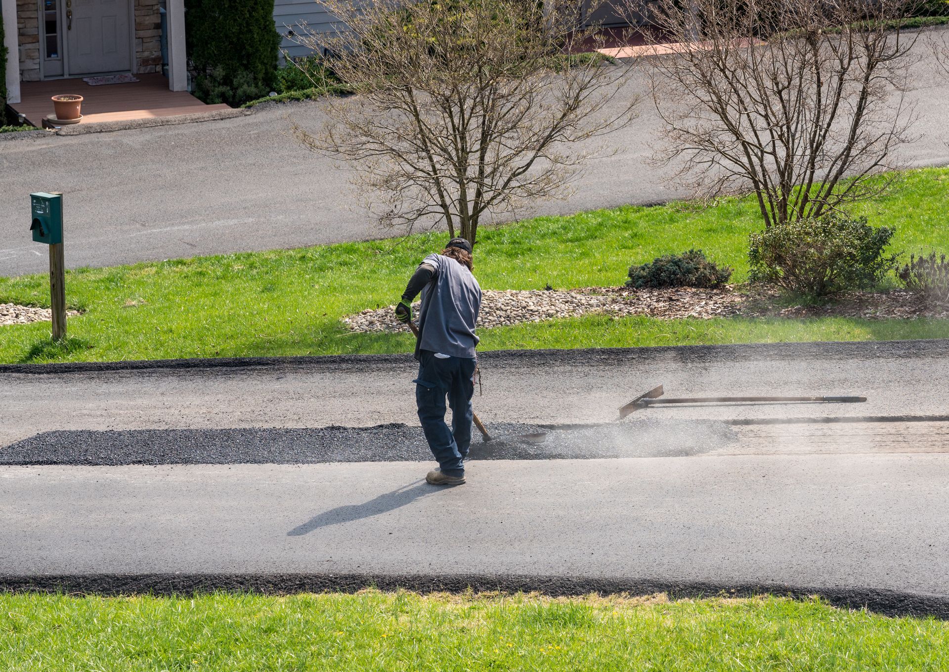 Person paving a road with asphalt, using a rake-like tool, with trees and a house in the background.