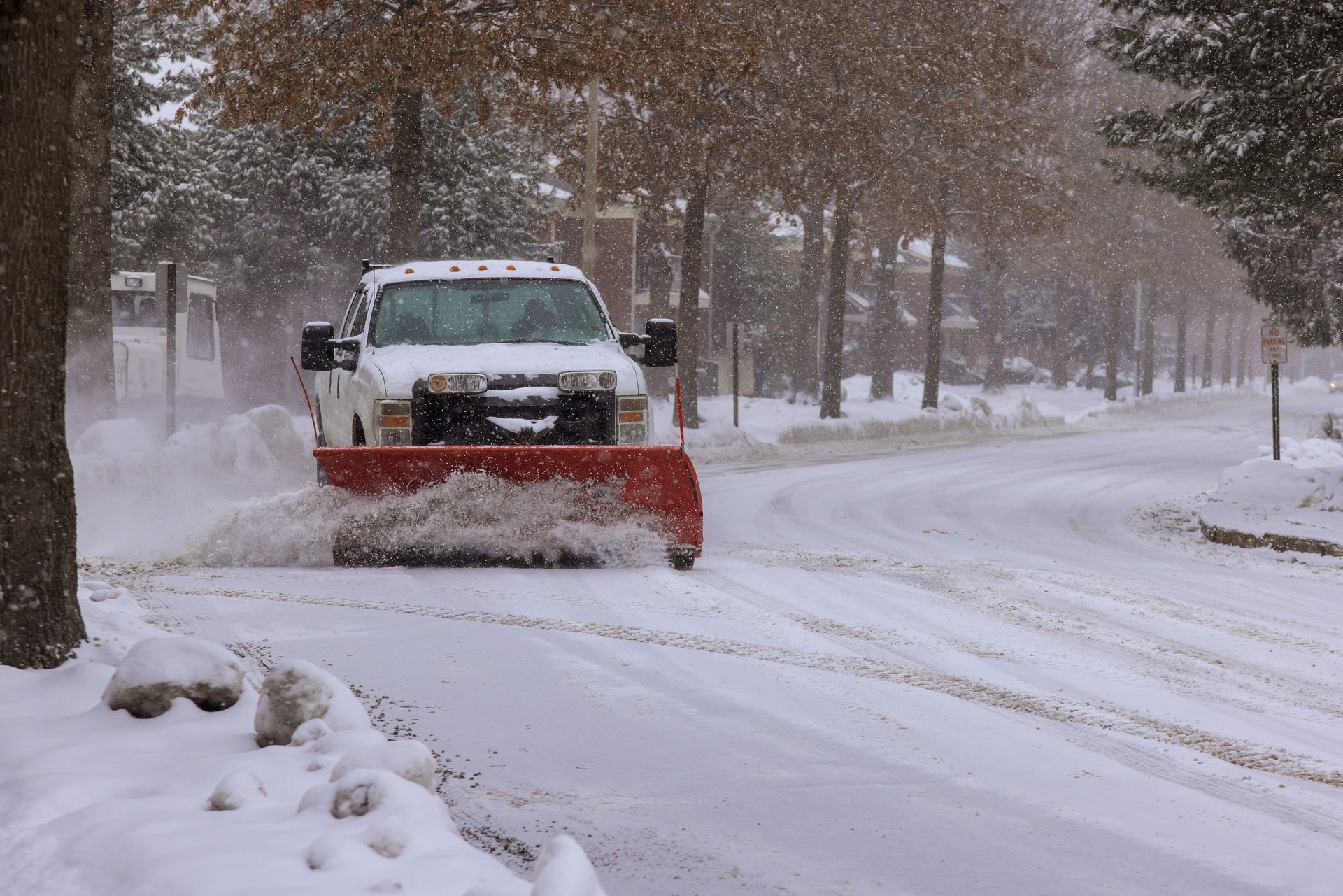 Snowplow clearing a snow-covered street during a snowfall. Red blade pushes snow to the side. Trees line the road.