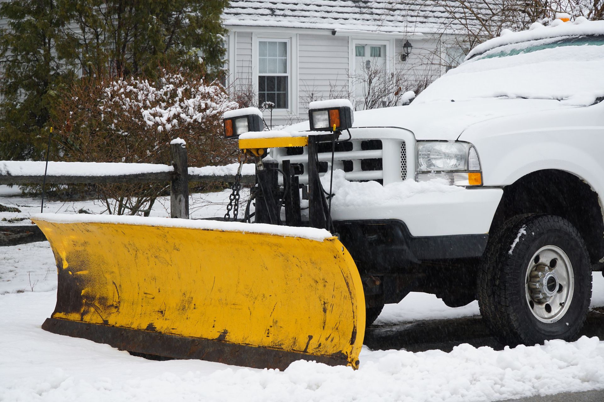 Yellow snowplow on a white truck clearing snow on a street in front of a house.