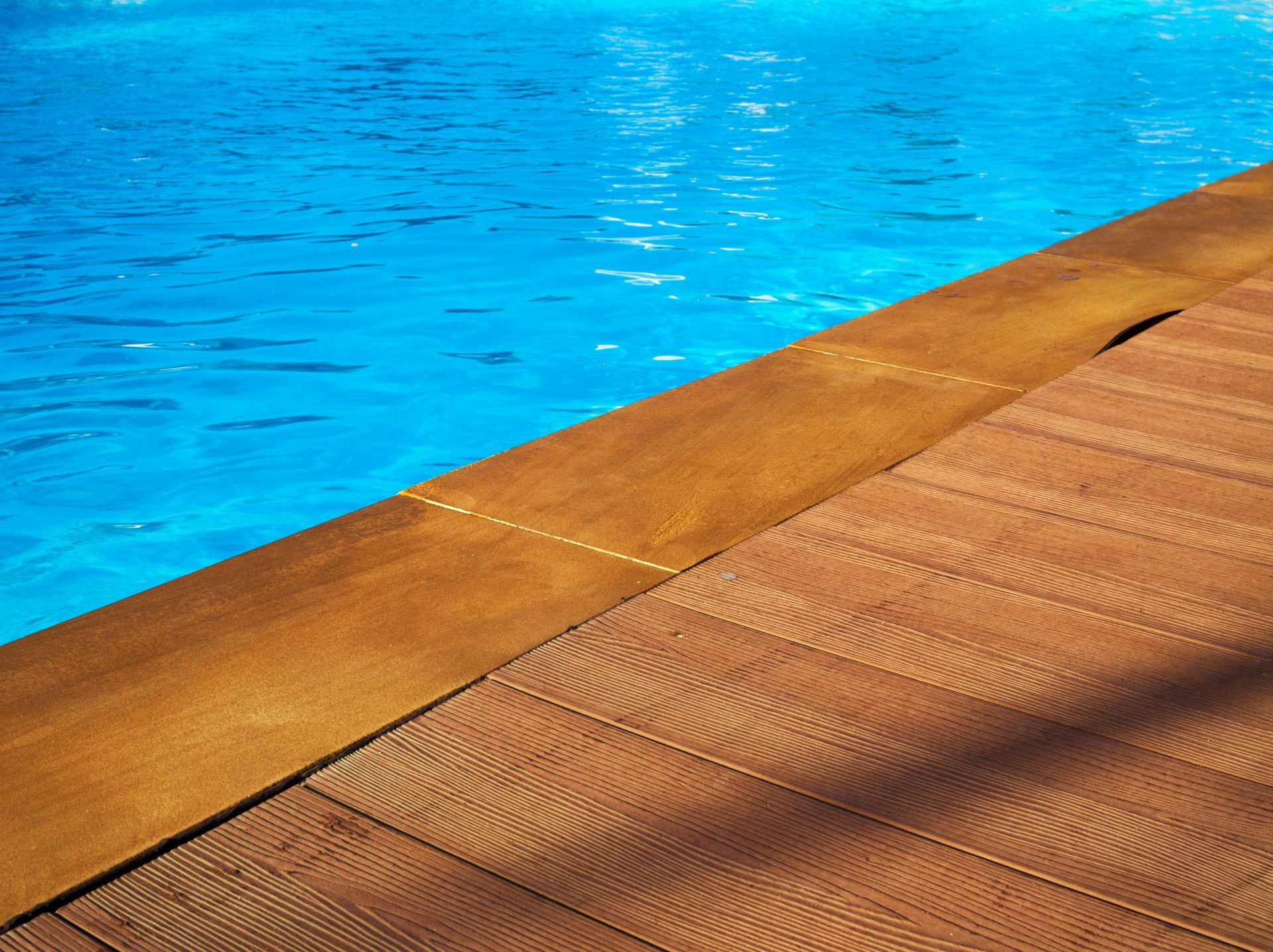 Poolside with wood-like decking, next to blue water, in bright sunlight.