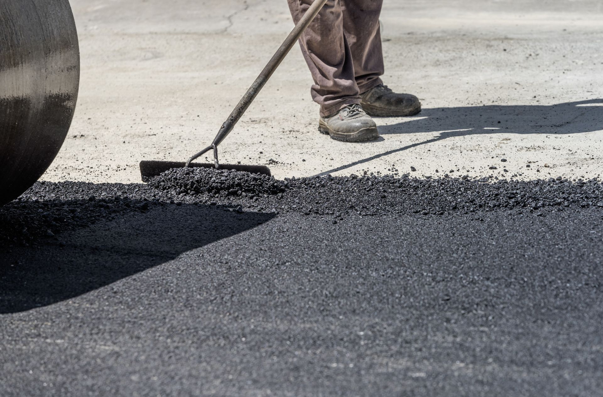 Worker smoothing fresh asphalt with a rake next to a road roller.