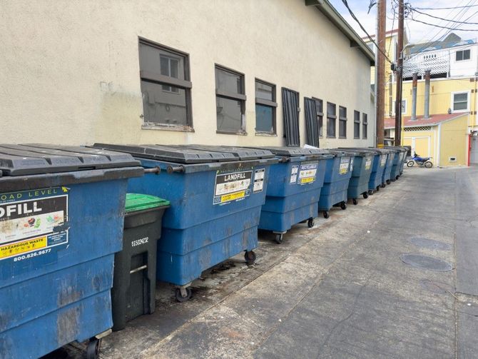 Row of blue trash bins lined along a narrow alley beside a beige building.
