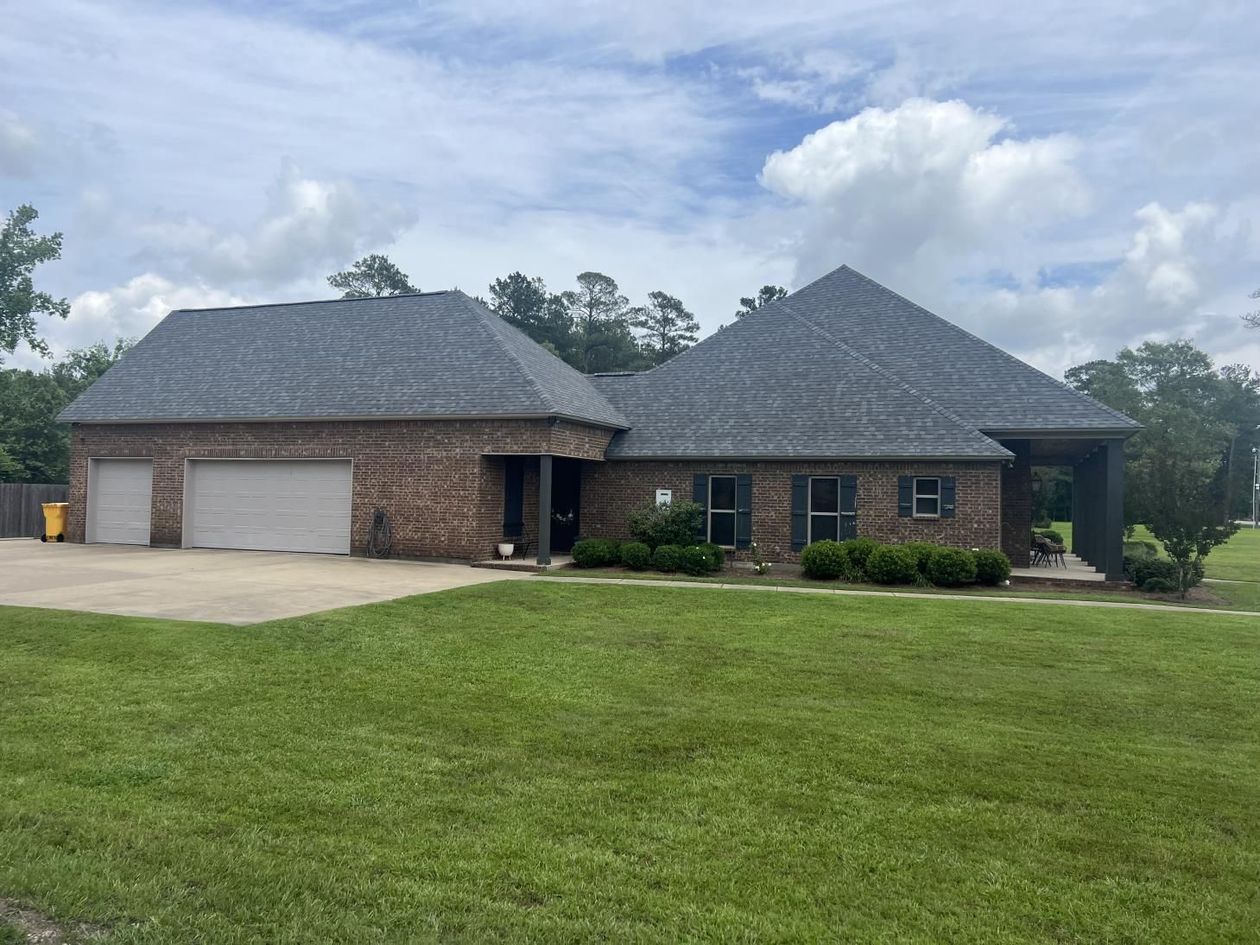 A large brick house with a gray roof is sitting on top of a lush green field.
