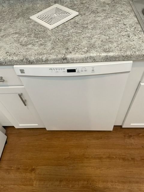 White Kenmore dishwasher installed under a granite countertop and above wooden flooring.