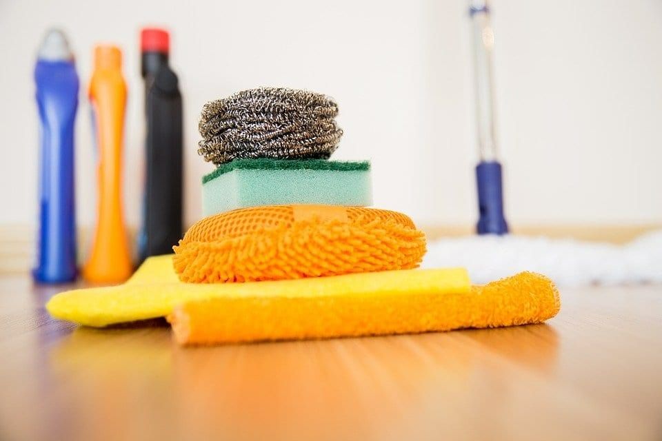 Cleaning supplies on a wooden floor: bottles of cleaner, sponges, scrubbers, and a mop.