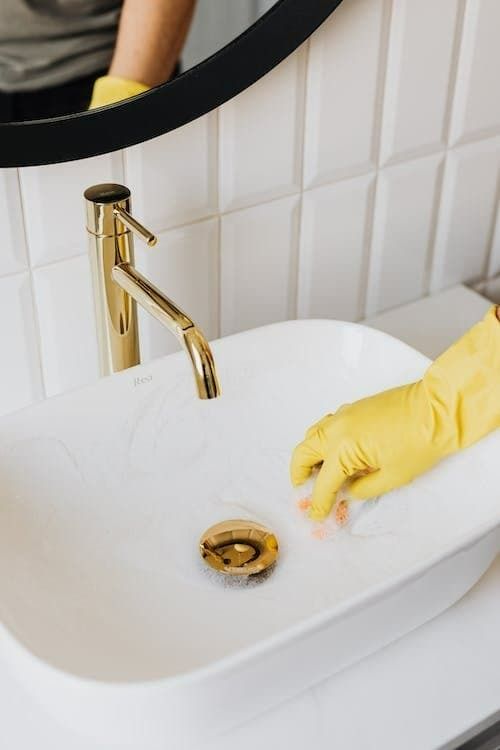 Person wearing yellow gloves cleans a white bathroom sink with a gold faucet.