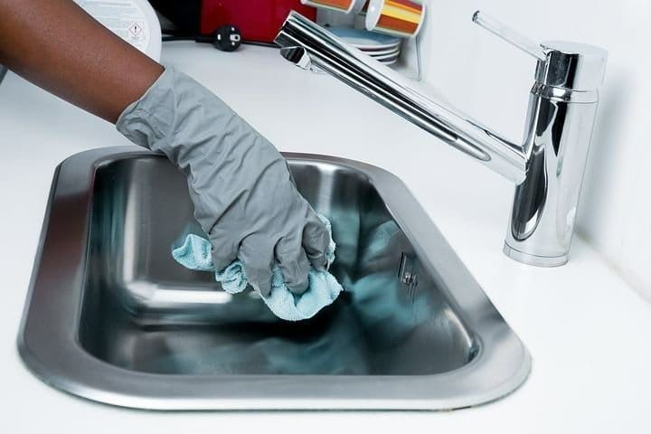 Person in a grey glove cleaning a stainless steel kitchen sink with a blue cloth.