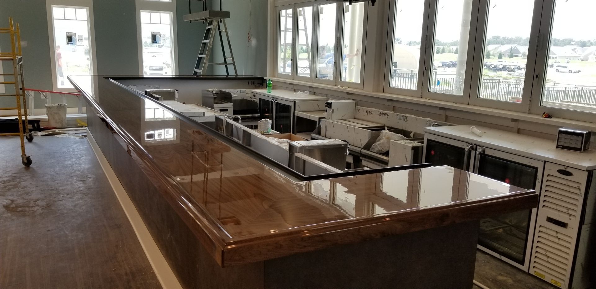 A long wooden bar with shiny countertop in a well-lit room, likely under construction. The bar has a stainless steel area for sinks and refrigeration.